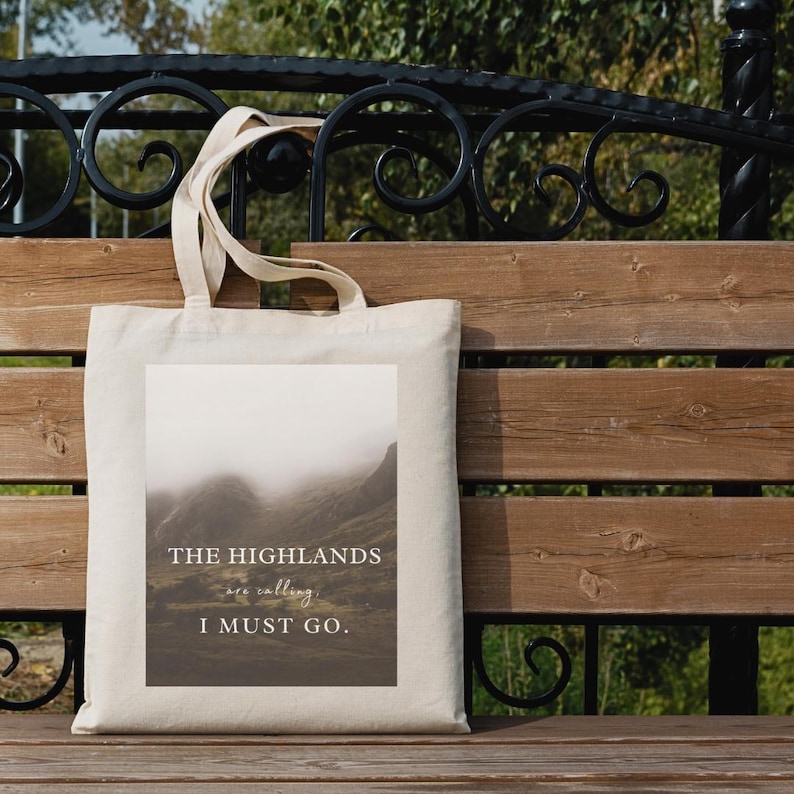 May include: A beige canvas tote bag with a rectangular image of a misty mountain landscape. The text "THE HIGHLANDS are calling, I MUST GO." is printed on the image in a serif font. The bag has two handles and is resting on a wooden bench.
