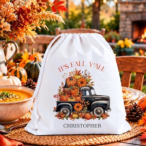 May include: A white drawstring bag featuring a graphic of a black vintage pickup truck overflowing with orange pumpkins and autumn leaves. The text "IT'S FALL Y'ALL" is printed in red above the truck, and the name "CHRISTOPHER" is printed in black below.