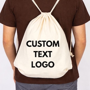 Op de afbeelding: Een crèmekleurige rugzak met trekkoord met de woorden "CUSTOM TEXT LOGO" in vette zwarte letters. De tas is gemaakt van een zachte, natuurlijke stof en heeft witte touwriemen.