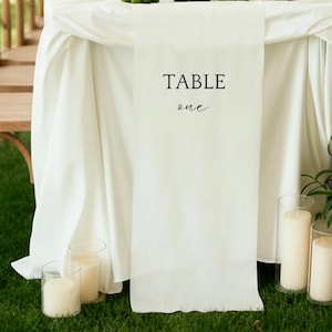 Può includere: Un runner da tavolo bianco con le parole "TABLE one" stampate in nero. Il runner è drappeggiato su una tovaglia bianca. Diverse candele in portacandele di vetro sono sull'erba sotto il tavolo.