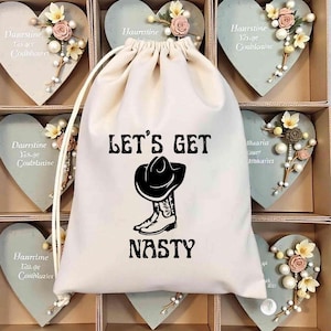 Puede incluir: Una bolsa blanca con cordón y la frase "LET'S GET NASTY" con un dibujo de sombrero y bota de vaquero. La bolsa está rodeada de decoraciones en forma de corazón con detalles florales, ideal para regalo.