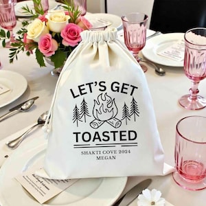 Puede incluir: Una bolsa blanca con cordón con el texto "LET'S GET TOASTED" sobre un gráfico de una fogata y árboles. La bolsa también incluye el texto "SHAKTI COVE 2024 MEGAN". La bolsa está sobre una mesa.