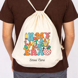 May include: A cream-colored drawstring backpack with the text "In My Last Day Of School Era" in colorful, retro-style lettering. The design includes a pencil, stars, a smiley face, and a checkered apple. The words "School Name" are below the main text.