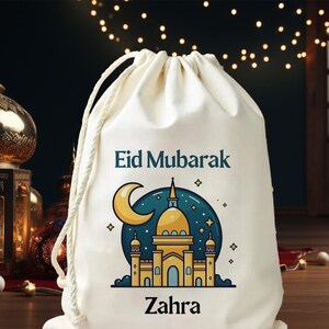 Puede incluir: Bolsa con cordón de color crema con el texto "Eid Mubarak" y el nombre "Zahra" en azul oscuro. La bolsa presenta una colorida ilustración de una mezquita con una luna creciente y estrellas, ideal para regalar.