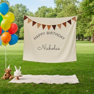 Può includere: Uno striscione color crema con la scritta "HAPPY BIRTHDAY Nicholas" e bandierine triangolari marroni. Un mazzo di palloncini colorati, un orsacchiotto, una piccola torta e un tappeto bianco sono sull'erba.