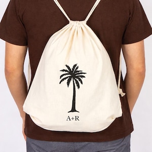 May include: A cream-colored drawstring backpack with a black palm tree graphic and the initials "A+R" printed below. The bag has white rope straps and is worn on the back.