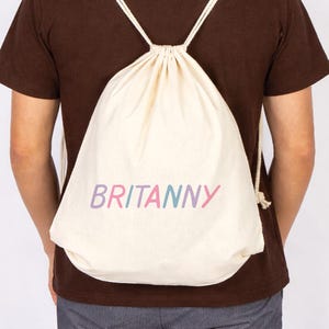 May include: A cream-colored drawstring backpack with the name "BRITANNY" printed in a colorful, pastel font. The bag is made of a soft, natural fabric and has white rope straps. The person is wearing a brown shirt.
