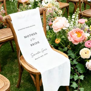 May include: A white fabric chair sash with the text "RESERVED FROM HEAVEN", "MOTHER of the BRIDE", "Samantha Grace", and "March 14th, 2026" in black. The sash is draped over a wooden chair, with flowers in the background.