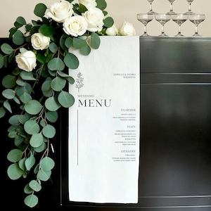 Può includere: Un menu di nozze bianco con testo nero, con i nomi "Estelle & Pedro" e un elenco di piatti. Il menu è decorato con un motivo floreale e poggia su una superficie scura, adornata da rose bianche ed eucalipto.