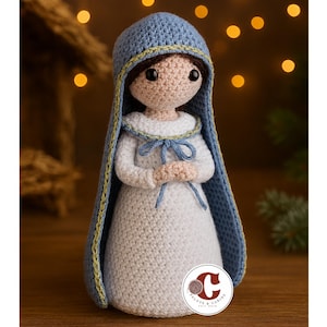 Puede incluir: Muñeca de ganchillo de una figura religiosa, vestida con un vestido blanco y un tocado azul con ribete dorado. La muñeca tiene una expresión neutra y las manos juntas. El fondo es borroso, con una iluminación suave y cálida.