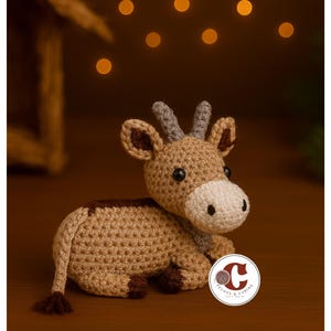 Può includere: Una statuetta di cervo all'uncinetto nei toni del marrone, beige e crema. Il cervo amigurumi fatto a mano ha piccole corna, un muso bianco e occhi neri. Il cervo è sdraiato su una superficie di legno.