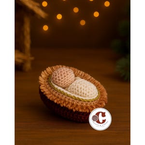 Puede incluir: Un belén hecho a mano con ganchillo. La escena presenta una cuna marrón con flecos marrón claro y dos figuras de ganchillo, una beige y otra rosa. La escena está sobre una superficie de madera con un fondo borroso.