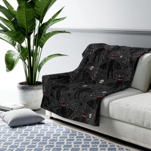 May include: A black throw blanket featuring a repeating pattern of winged moth-like creatures with bright red eyes, interspersed with small white skulls, stars, and crescent moons. The fabric has a soft texture and a dark, detailed graphic print.