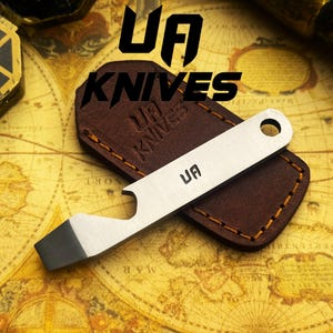 May include: A silver bottle opener with a dark gray tip and the letters "UA" is displayed on a brown leather sheath. The words "UA KNIVES" are printed in black above the sheath. The background is a vintage map.