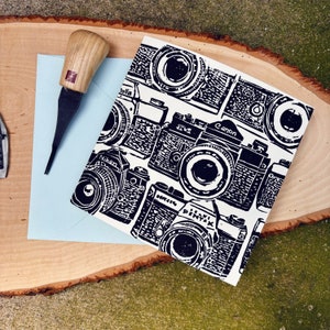 May include: A white card with black and white illustrations of vintage cameras. The cameras are arranged in a pattern and include brands like Nikon, Canon, and Pentax.