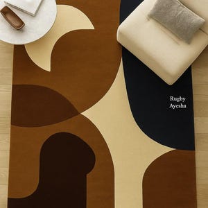 Op de afbeelding: Een rechthoekig vloerkleed met een abstract ontwerp in bruin-, beige- en marineblauwtinten. Het kleed heeft gebogen vormen en een gestructureerd oppervlak. De tekst "Rugby Ayesha" is zichtbaar op het kleed.