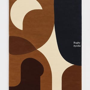 Op de afbeelding: Rechthoekig tapijt met een abstract ontwerp in bruin-, cr&egrave;me- en marineblauwtinten. Het tapijt heeft gebogen vormen en een halve maan ontwerp. De tekst "Rugby Ayesha" staat in de rechter benedenhoek.