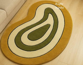 May include: An oval-shaped rug with a retro design. The rug features concentric bands of mustard yellow, cream, olive green, and black. The text "Rugby Ayesha" is visible on the rug.