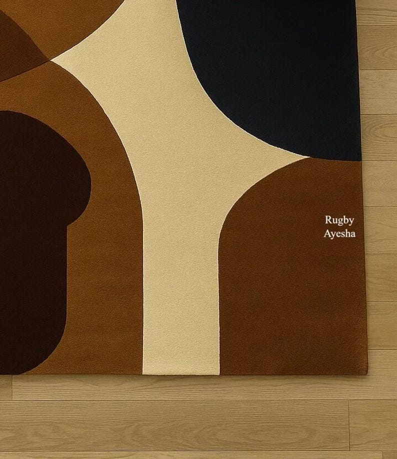 Op de afbeelding: Een modern vloerkleed met een abstract geometrisch ontwerp. Het heeft grote, gebogen vormen in bruin, beige en marineblauw. De tekst "Rugby Ayesha" is op het vloerkleed gedrukt.