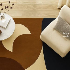 Op de afbeelding: Een modern vloerkleed met een geometrisch ontwerp in bruin, beige en zwart. Een kleine, ronde witte tafel met een vaas en een boek staat op de houten vloer. Een beige fauteuil met een grijs kussen is ook zichtbaar.