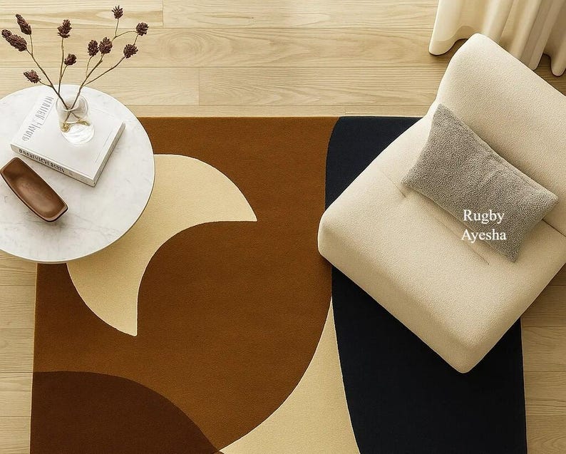 Op de afbeelding: Een modern vloerkleed met een geometrisch ontwerp in bruin, beige en zwart. Een kleine, ronde witte tafel met een vaas en een boek staat op de houten vloer. Een beige fauteuil met een grijs kussen is ook zichtbaar.