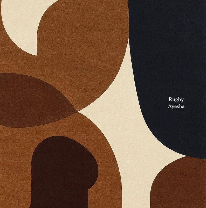 Op de afbeelding: Een modern vloerkleed met een abstract ontwerp. Het vloerkleed heeft grote, overlappende vormen in bruin-, cr&egrave;me- en marineblauwtinten. De tekst "Rugby Ayesha" staat in de rechter benedenhoek. Het vloerkleed is waarschijnlijk gemaakt van zacht materiaal.