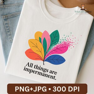 Pode incluir: Camiseta branca com um design floral colorido com folhas laranja, amarelas, rosa, verdes e azuis. O texto "All things are impermanent" está impresso abaixo. A camiseta está dobrada sobre jeans azuis.