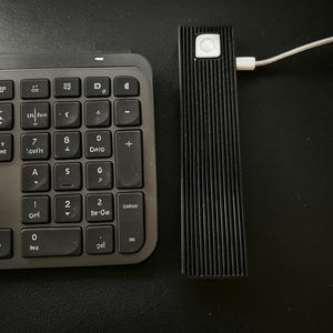 Può includere: Un dispositivo elettronico rettangolare nero con superficie scanalata e un pulsante circolare bianco sulla parte superiore, collegato a un cavo di ricarica bianco, posizionato accanto a una tastiera per computer nera con tasti grigio scuro.