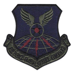 May include: A navy blue embroidered patch with a shield shape. The patch features a design with wings, a star, and a globe with red lightning bolts. The bottom of the patch has text in a curved banner.