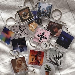 May include: A collection of clear acrylic keychains with various images, including cityscapes, portraits, and abstract designs. Each keychain has a silver metal ring. Some keychains feature text, and the overall aesthetic is modern and artistic.