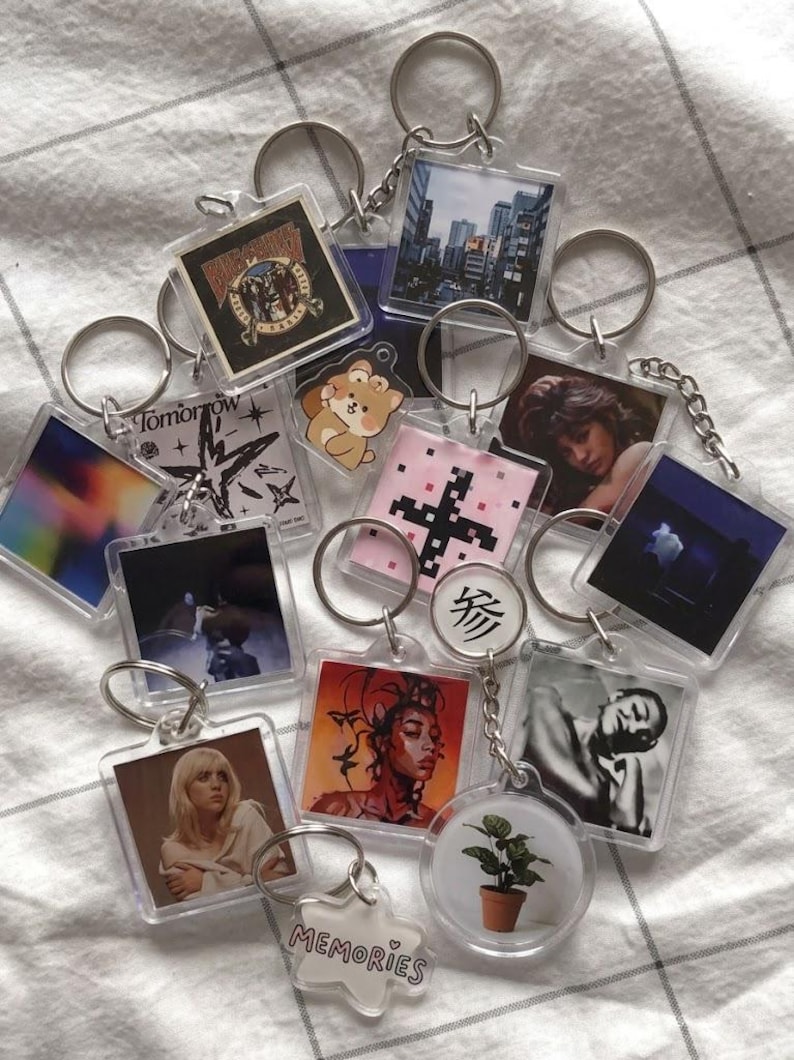 May include: A collection of clear acrylic keychains with various images, including cityscapes, portraits, and abstract designs. Each keychain has a silver metal ring. Some keychains feature text, and the overall aesthetic is modern and artistic.