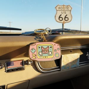 May include: A gold-toned metal keychain shaped like a handheld video game console in light pink. The screen features a pixelated cat character with a crescent moon symbol on its forehead, surrounded by small white wings and heart icons.
