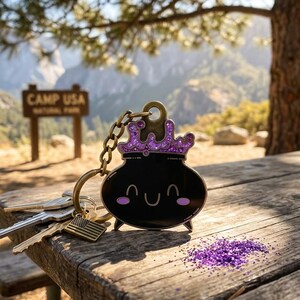 May include: A black cauldron keychain with a purple jeweled crown and a smiling face. The keychain is attached to a chain and a small American flag charm. Keys are on a wooden table with purple glitter.
