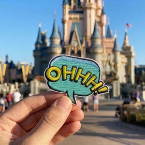 May include: A teal speech bubble patch with the word "OHHH!" in yellow, outlined in black. The patch is held in front of a castle with blue spires and a blue sky.
