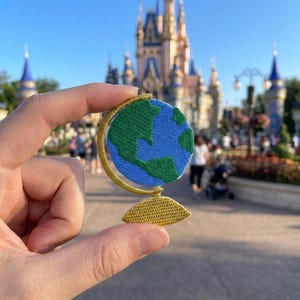 May include: Embroidered globe patch featuring a blue and green world map with gold-colored accents. The patch is held up in front of a castle and park setting.