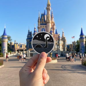 May include: A round, black and white patch featuring a stylized Loch Ness Monster design. The patch shows the monster in the water under a night sky with stars and a moon. The patch is held in front of a castle.
