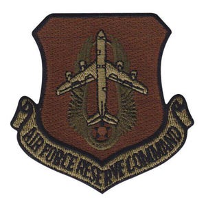 May include: A brown and gold embroidered patch featuring an airplane design at the center. The patch has a shield shape with a banner at the bottom that reads "AIR FORCE RESERVE COMMAND". The patch is likely a military insignia.