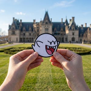 May include: A white, circular patch featuring a cartoon ghost with a wide, toothy grin and a pink tongue. The ghost has black eyes and a black outline. The patch is held in front of a large building and green lawn.