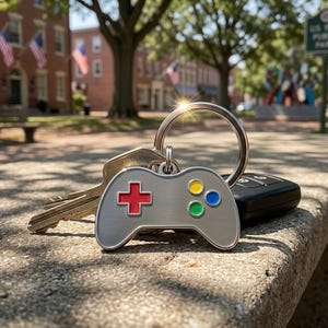 May include: A silver-colored keychain with a video game controller design. The controller has a red plus sign, and yellow, blue, and green buttons. The keychain is attached to keys and a car remote. The background shows a park setting.