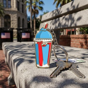 May include: A colorful keychain featuring a cartoon-style slushy drink design. The keychain has a blue, red, and white color scheme, with a gold-colored border. The keychain is attached to a silver chain and key ring, with two keys resting nearby.
