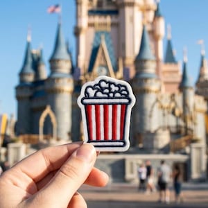 May include: A popcorn patch with a red and white striped bucket design, filled with white popcorn. The patch is held in front of a castle with blue turrets and a blue sky.
