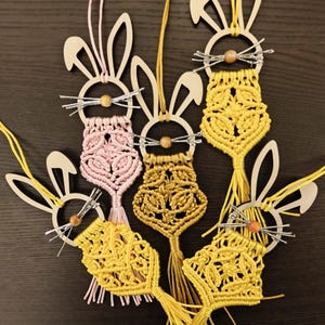 Könnte beinhalten: Macramé-Hasenornamente in Gelb, Rosa und Gold. Jeder Hase hat einen Holzkopf mit ausgeschnittenen Ohren, eine Holzkugel-Nase und Schnurrhaare aus Silberdraht. Die Makramee-Körper weisen aufwendige Knotenmuster auf.