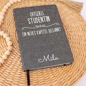 Könnte beinhalten: Ein dunkelgraues, stoffbezogenes Notizbuch mit weißem Text "OFFIZIELL STUDENTIN EIN NEUES KAPITEL BEGINNT" und dem Namen "Mila" darunter. Eine lange Kette aus hellen Holzperlen mit einer Jutequaste ist um das Notizbuch herum drapiert.