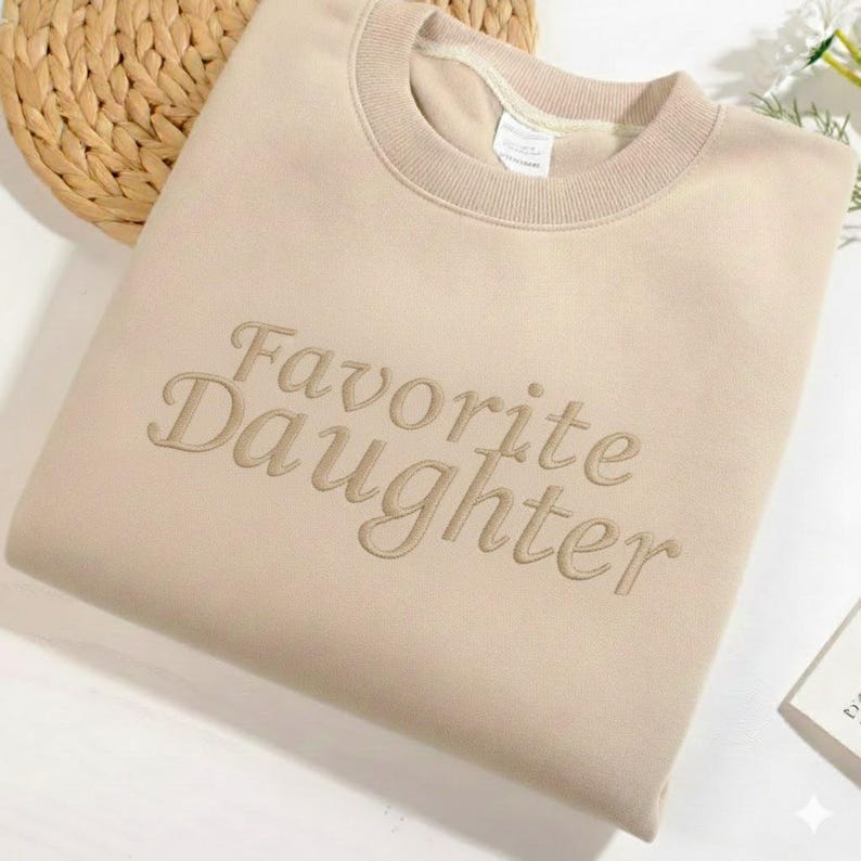 May include: A beige sweatshirt with the embroidered text "Favorite Daughter" in a matching tone. The crew neck sweatshirt is folded neatly on a white surface, with a woven basket in the background.