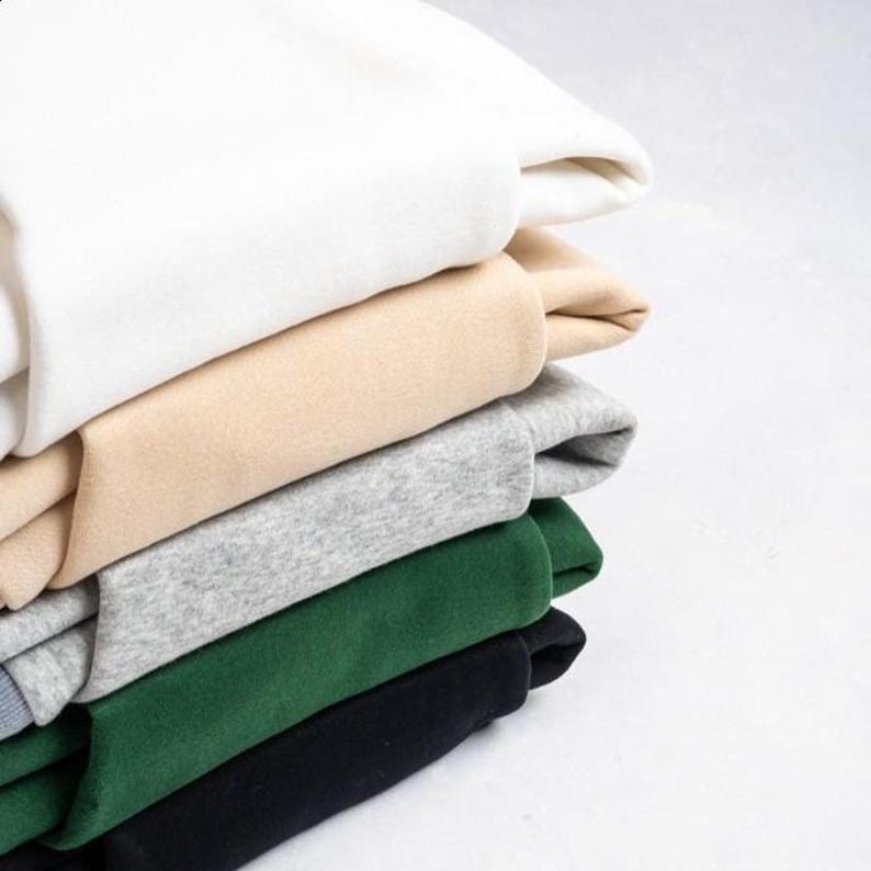 May include: A stack of folded sweatshirts in various colors: white, beige, gray, forest green, and black. The sweatshirts are neatly arranged, showcasing the different hues and textures of the fabric. The image is well-lit, highlighting the soft folds of the clothing.