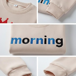 May include: Beige sweatshirt with the word "morning" embroidered in blue and black. The sweatshirt has a crew neck and ribbed cuffs. A red crown is embroidered on the upper left side. The sleeves are long.