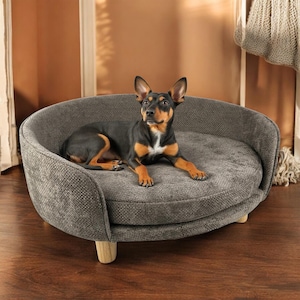 Puede incluir: Una cama para perros gris de forma ovalada con respaldo redondeado y patas de madera. Un perro negro y marrón está acostado cómodamente en la cama. La cama tiene una tela texturizada y un diseño moderno.
