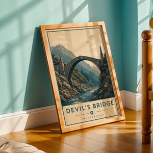 May include: A framed print with a vintage travel poster design. The artwork shows the Devil's Bridge in Switzerland, a stone arch bridge over a river, with mountains in the background. The poster has a light brown border and the text "DEVIL'S BRIDGE SWITZERLAND".