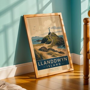 May include: A framed print featuring a vintage travel poster design. The artwork depicts Llanddwyn Island, Anglesey, North Wales, with a lighthouse atop a rocky hill, a winding path, and the sea. The frame is made of light-colored wood.