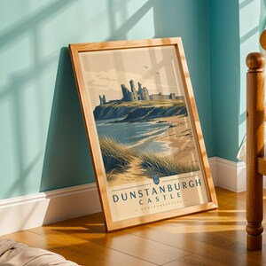 Op de afbeelding: Een ingelijste prent met Dunstanburgh Castle in Northumberland. De afbeelding toont de kasteelruïnes op een klif met uitzicht op een strand en de zee. De prent heeft een lichtbruine houten lijst en de tekst "DUNSTANBURGH CASTLE - NORTHUMBERLAND".